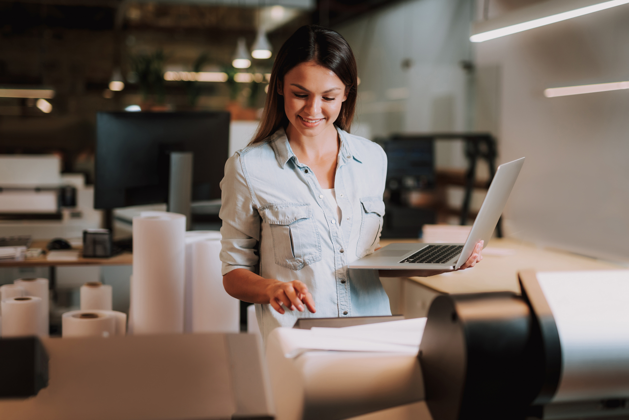 Naine seisab kontorikeskkonnas, kasutab suurt printerit ja hoiab teises käes sülearvutit.